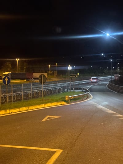 This image captures a nighttime view of a road interchange or access point within the city of Tortoreto, Italy. In the foreground, an asphalt surface with a white triangular yield marking and yellow boundary lines curves to the right. A silver metal guardrail follows this curve. To the left, a grassy median separates the foreground road from a lower, parallel section, likely a railway or service road, secured by a metal fence. A white semi-truck with a dark red trailer is positioned on this parallel section to the left.

In the mid-ground, a multi-lane highway is visible, where a white van, exhibiting illuminated rear and possibly roof-mounted lights, is observed driving away from the viewer. Streetlights line the highway and surrounding areas, casting light and generating prominent lens flares, particularly in the upper right portion of the frame. Road signage includes a green sign with "ANCONA" and an arrow pointing right, a blue circular sign with an upward white arrow, a black circular prohibitory sign, and a red and white triangular yield sign on the far right. The sky is dark and clear. No people are discernible in the scene.
