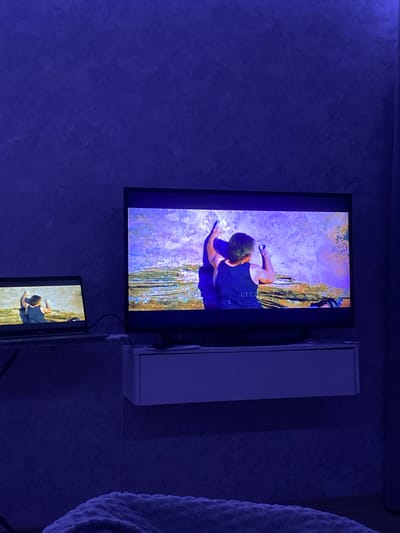 The image shows a blue-lit interior with two vertically stacked screens displaying a rock climber. The larger, upper screen shows the climber at the top of a rock face or cliff, while the smaller, lower screen offers a closer view of the climber's upper body. A white cabinet is adjacent to the upper screen. The location of the image is Зубово, Russia.