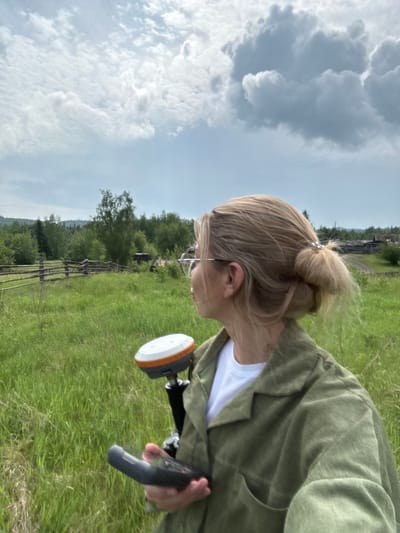 A woman with blonde hair tied in a bun is standing in a grassy field, holding a surveying device. She is wearing glasses and a green jacket over a white t-shirt. The background shows a wooden fence, trees, and a few rustic buildings. The sky is cloudy. The location is identified as Badarminsk, Russia.