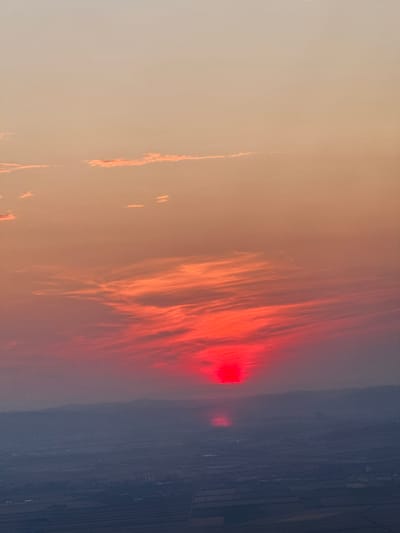 This image depicts a vibrant sunset over a landscape that includes a hint of a city or town, likely Enzersdorf an der Fischa, Austria, given the context. The sky is filled with streaks of intense red and orange clouds, illuminated by a bright red sun partially obscured by atmospheric haze. The sun itself appears as a luminous disc, casting a fiery glow. Below the sky, a silhouetted mountain range is visible, with a darker, hazy expanse stretching out in front of it. Within this lower expanse, faint signs of human settlement can be discerned, appearing as muted lights or structures against the darkening landscape. The overall impression is one of a dramatic and colorful natural phenomenon occurring at dusk.