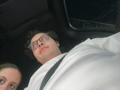 A photograph of two individuals, a man and a woman, is presented from an atypical, near-overhead angle. The man, located towards the bottom left, is wearing glasses and a white shirt. The woman is partially visible in the upper right corner, only her eyes showing. The background appears to be the interior of a vehicle, judging by the outline of the car's roof and window visible. The overall lighting is dim, giving the scene a somewhat shadowy atmosphere.