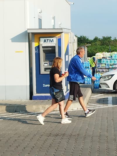 This image depicts an outdoor scene, likely at a service station or convenience store, in Cupele, Romania. In the foreground, two individuals are walking. A woman in a black t-shirt and denim shorts, with shoulder-length blonde hair, is holding a red cup. A man in a blue jacket and black shorts walks beside her. Behind them, a person wearing a high-visibility vest is visible near stacks of bottled water. An ATM is situated against a white building. The ground is paved with interlocking grey bricks, and a white car is partially visible on the right side of the frame. The scene is illuminated by natural daylight, suggesting it is daytime.