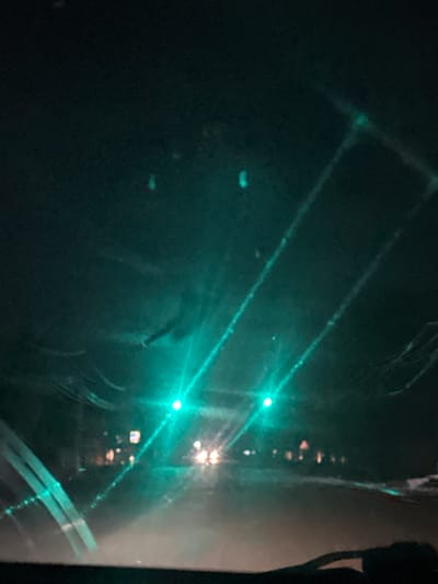The image depicts a nighttime scene from inside a vehicle, looking forward through a wet windshield. Bright green lights from traffic signals are prominent, casting a teal hue over the surroundings. Streaks of light, possibly from headlights or reflections, traverse the frame. In the distance, blurred lights suggest buildings or other vehicles. The road surface is obscured by mist or fog, contributing to the low visibility. The overall impression is one of driving in challenging conditions, likely during a rainy or foggy night in an urban or semi-urban environment. The location is identified as Кызыл-Кыргызстан, Kyrgyzstan.