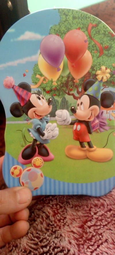The image depicts a close-up, top-down view of a birthday-themed cutout featuring Mickey and Minnie Mouse. They are standing in a grassy outdoor setting with balloons and a tree in the background. Minnie Mouse is on the left, wearing a party hat and blue dress, and Mickey Mouse is on the right, wearing a red shirt and party hat. They appear to be interacting, with Mickey extending a hand. In the foreground, there are round sticker-like objects with concentric circles and dots, resembling a target or a Mickey Mouse ear motif. The cutout is positioned on a textured, reddish-brown surface, possibly a blanket or rug. The image is taken indoors, with a dark, out-of-focus area in the top left corner suggesting a person holding the cutout or part of the indoor environment. The location in Marhamat, Uzbekistan, is not depicted in the image itself and is a separate piece of information provided in the prompt.
