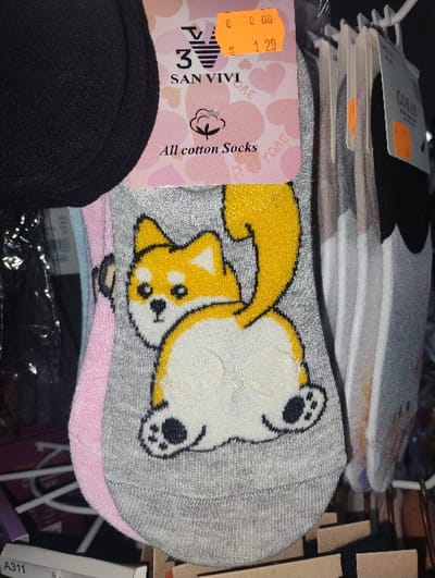 A pair of grey ankle socks, adorned with a cartoon depiction of a yellow and white dog, possibly a Shiba Inu or Corgi, facing away and looking over its left shoulder, is the central focus. The socks are affixed to a pink and white cardboard label that features the brand "SAN VIVI" with a stylized "3" logo, the text "All cotton Socks," and a background pattern of hearts with the repeated word "ТОЛЕ". An orange price sticker on the label displays "€ 0.86" and "1.29." Partially visible on the upper left is a section of a black fabric item. To the right, several other pairs of socks, packaged in clear plastic, hang on a white plastic retail display hook; some of these also bear orange price tags. A white label with "A311" is visible in the bottom left foreground. The scene depicts a retail display in what appears to be an indoor commercial setting within the city of Byal Izvor, Bulgaria.