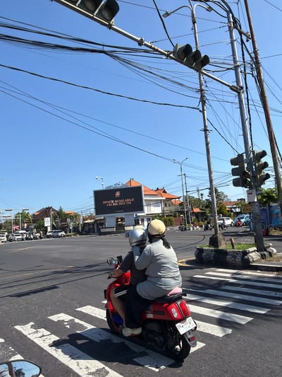 The image captures a street scene in Denpasar, Indonesia, with a bright blue sky overhead and a complex network of overhead electrical wires crisscrossing the frame. In the foreground, a red scooter carrying two people is stopped at a pedestrian crossing. The person in the back, wearing a grey top and dark pants, is leaning forward. The person in front, wearing a white helmet, is looking forward. Both are wearing helmets. The scooter has a license plate that reads "DK 3566 ADP".
Further in the background, traffic is visible at an intersection. Cars and motorcycles are present, and a traffic light hangs above the intersection. A large digital billboard on the right side of the road displays "SPACE AVAILABLE" and a phone number. Buildings with orange-tiled roofs are visible behind the billboard and along the street, characteristic of Indonesian architecture. The overall environment appears to be a busy urban intersection.