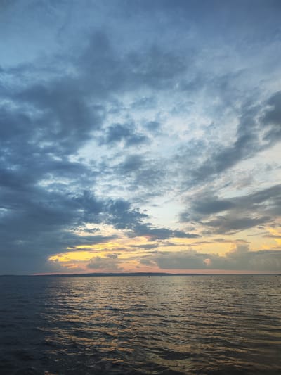 The image shows a vast body of water under a cloudy sky, likely a lake or sea. The water is dark blue with ripples reflecting the light from the sky. The sky is a mix of dark blue and gray clouds, with patches of yellow and orange light breaking through near the horizon, indicating either sunrise or sunset. A faint, dark line representing land is visible in the distance, with a few small, dark shapes that might be boats or distant figures. There are no discernible people or specific structures in the foreground or midground. The scene appears to be a natural landscape.