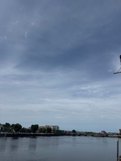 This is a photograph of the River Shannon in Limerick, Ireland, looking east. The sky is overcast with patches of white clouds. The river is calm and dark. On the far bank, a promenade is lined with trees and buildings, including some modern apartment blocks and older brick structures. Several bridges cross the river in the distance. A tall, dark structure, possibly a crane or part of a building, is visible in the upper right corner of the frame. There are no people visible in the image.