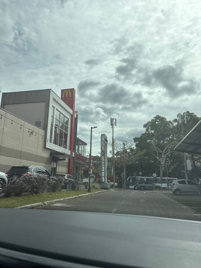 The image shows a view from the driver's seat of a car looking down a parking lot. The lot is mostly empty, except for a few parked cars and a bus parked toward the back.  The parking lot is located next to a large McDonald's building with a red awning.  There are trees on the right side of the parking lot, and the building is partially obscured by their foliage.  The sky is cloudy and overcast.  The location is San Rafael, Costa Rica, at coordinates 9.937, -84.152.