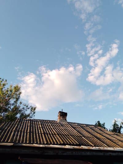 The image presents an upward view of an aged, corrugated tile roof and a brick chimney, set against a bright blue sky dotted with scattered white clouds. The roof, which appears to be made of asbestos cement or a similar material, shows distinct signs of weathering and discoloration, with dark streaks and some lighter, possibly moss-covered patches along its ridges. A rustic, dark-colored brick chimney rises from the center of the roof's peak.
To the left, lush green foliage from a tree extends into the frame, its leaves catching some sunlight. On the right, the top branches of another tree are partially visible against the sky. The lighting suggests either early morning or late afternoon, with a low sun angle casting warm, golden light on the prominent white clouds and highlighting the texture of the roof tiles, creating clear shadows within the corrugations. The sky is predominantly clear and bright.
There are no people or specific activities depicted. The scene conveys a sense of a quiet, sunny day in a residential or semi-rural setting, consistent with the provided location context of San Martín de Porres, Peru. No text is visible in the image.