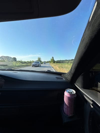 An image, originally rotated ninety degrees clockwise, captures a view from inside a moving vehicle on a sunny day. The perspective looks forward through the windshield, revealing a clear blue sky and a road stretching into the distance. The scene suggests a rural or semi-rural setting, with green grassy fields and trees lining the roadside under a bright sun, indicative of morning or early afternoon.

In the foreground, the dark interior of the car is visible, including the A-pillar on the right and part of the dashboard or door panel. A pink aluminum beverage can is snugly placed in a cupholder or storage slot within the dashboard area to the right. Text on the can is partially legible, clearly showing "AROMATIZED" and likely "CARBONATED" below it, though the full brand name is not discernible.

On the road ahead, a dark-colored car, possibly black or dark grey, is clearly visible, with its rear facing the viewer. Further down the road, another lighter-colored vehicle can be seen. The road appears to be a single-lane carriageway in each direction. The overall impression is one of travel through a pleasant, open landscape, with the provided context placing the location in Krivodol, Bulgaria.
