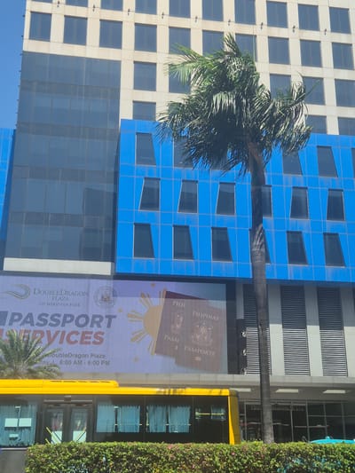 The image shows the DoubleDragon Plaza in Pasay, Philippines. The building is partially visible, showcasing its modern architectural design with a mix of white and blue panels. A tall palm tree is positioned in front of the building, partially obscuring the facade. A large advertisement banner for "Passport Services" is displayed on the building's exterior. A yellow bus is parked next to the building, partially obscured by a dense row of bushes.