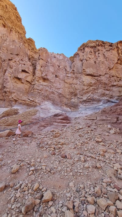 A young girl in a pink dress and hat stands on a rocky, uneven terrain at the base of a steep rock cliff face. The cliff, composed of reddish-brown stone, is marked with a series of metal bars that form a rudimentary ladder system ascending the rock face. The sky is visible above the cliff. This scene is located in the city of Hevel Eilot Regional Council, Israel.
