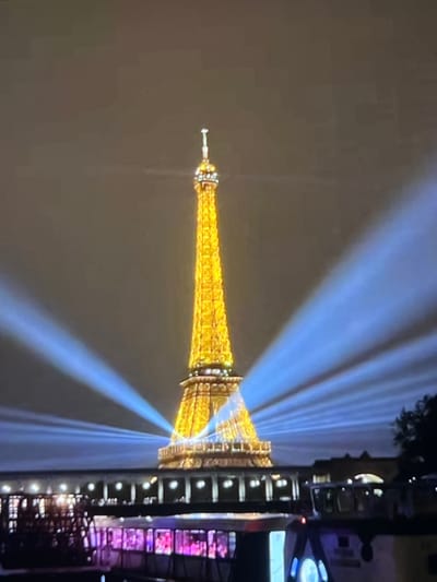 The image shows the Eiffel Tower illuminated with golden lights at night, with bright blue beams of light emanating from its base and reaching towards the sky. The tower is situated in the city of Paris, France, not Montfermeil. In the foreground, there is a body of water with several boats, one of which has colorful lights. A structure resembling a bridge or promenade runs along the water's edge in front of the tower, with lights illuminating it. The sky is dark and overcast.