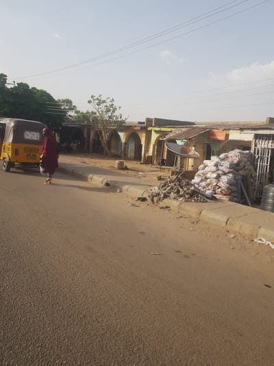 A medium shot captures a street scene in Dutsinma A, Nigeria, featuring a brown and yellow Bajaj tricycle, commonly known as a "Keke," on the left side of the asphalt road. A woman wearing a red head covering and gown is positioned in front of the vehicle. Stacks of white sacks are piled on the side of the road near a multi-story building. The building has a faded yellow façade, a green arched colonnade, and a satellite dish attached to the upper part of the building. Wires are stretched across the sky on the left side of the image.