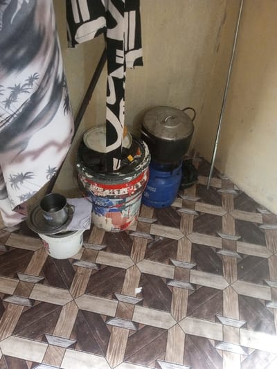 A corner of a room features a patterned floor and beige walls. A propane tank supports a cooking pot with a lid. Beside it, a stack of paint buckets serve as a table. On top of the buckets, a mug sits on a saucer and a cloth. Items of clothing are draped nearby, adding to the cluttered atmosphere. A thin, silver pole leans against the wall.