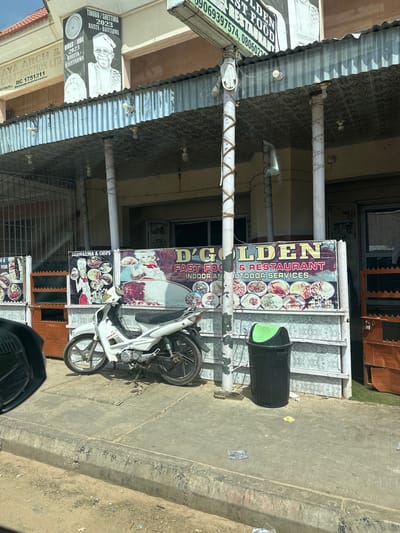 The image shows a storefront in Ajiwa, Nigeria, identified by a banner that reads "D'GOLDEN FAST FOOD & RESTAURANT INDOOR AND OUTDOOR SERVICES". To the left of this banner, there are two other smaller banners. One displays "SHAWARMA & CHIPS" with images of food and two cartoon figures in chef attire. Another banner to the far left appears to advertise "FISH". Above the storefront, a large poster features portraits of two individuals, identified as "TINUBU / SHETTIMA 2023" and "DIKKO / JOBE 2023 BACUTA - BACUTARWA". A white motorcycle is parked on the sidewalk in front of the storefront, leaning on its kickstand. A black trash can with a green lid is situated to the right of the main banner. The storefront itself has a corrugated metal roof and supporting columns wrapped with twine. The surrounding environment includes a concrete sidewalk and curb, with some scattered debris. The building behind the storefront is visible, with windows and signage. The lighting suggests it is daytime.