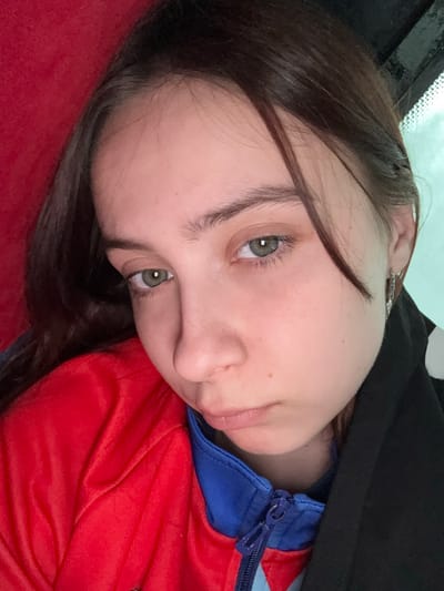 A close-up, angled, self-portrait depicts a young woman. Her face is pale with light brown hair. Her eyes are light green. She is wearing a red jacket with a blue collar. A black garment is draped over her shoulder. A vehicle windshield is visible at the top left of the frame. The setting is likely indoors, possibly inside a car. The location is unknown.