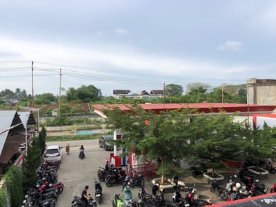 The image captures an outdoor scene at a gas station in Geulumpangsulu Timu, Indonesia. The time of day appears to be daytime, with overcast skies suggesting potential for rain or a generally cloudy atmosphere.
The foreground is dominated by numerous parked motorcycles, a common mode of transportation in the region. Several people are visible, some riding motorcycles and others standing or walking near them. Two individuals are walking near a white car parked in a lot. A person is seated on a motorcycle at the gas pumps, and another is standing near a motorcycle. Further back, several more individuals are either walking or on their bikes.
The gas station itself is characterized by a prominent red roof structure, typical of such facilities. Trees with lush green foliage are scattered throughout the scene, some planted in rectangular planters. In the background, buildings with red roofs and a large concrete structure are visible, along with a dense line of trees and vegetation, suggesting a semi-urban or suburban environment. Overhead power lines with wooden poles crisscross the sky, a common sight in many Indonesian locales.
A white car with an Indonesian license plate (likely reading "BL 1183 Y") is parked in the vicinity of the gas station, and a dark-colored SUV is positioned near the pumps. The overall impression is one of a bustling, everyday scene at a local service station. No specific emotions are discernible from the image; the focus is on the activities and objects present in the setting.