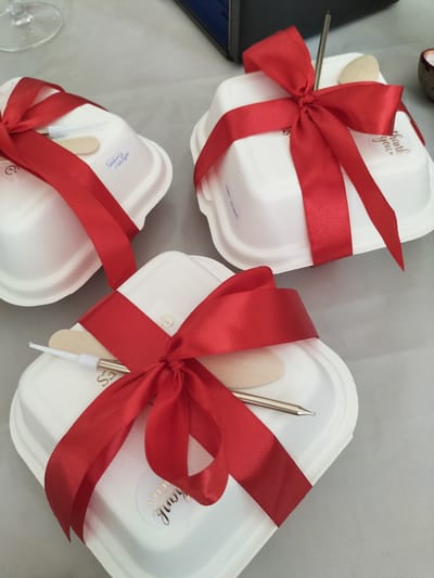 The image shows three white, heart-shaped takeaway containers, each tied with a bright red ribbon. These containers appear to be part of a catering or gift service. Each container holds a small wooden utensil and a metal skewer. One container has a sticker with "thank you" written in gold script. Another has "Белый натур" written on it, likely indicating the contents. The setting is a table covered with a light grey textured cloth, possibly for an event or gathering. A clear glass, possibly a wine glass, is visible in the upper left corner. The overall mood is festive and appreciative, suggested by the red ribbons and "thank you" message. The lighting is soft and diffused, indicating an indoor setting. The image is a close-up, focusing on the presentation of the takeaway items.