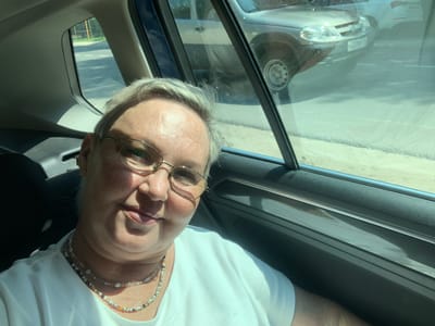 The image is a selfie taken from inside a car, looking out the window. A woman with light-colored hair and glasses is in the foreground, wearing a white t-shirt and two beaded necklaces. Her face is partially visible, and she is smiling slightly. Through the car window, another car is visible, along with a road and some greenery. The scene appears to be a sunny day.