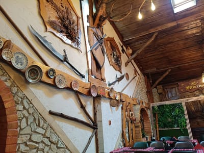 This image depicts the interior of a restaurant with a rustic and traditional theme, likely located in Câmpia Turzii, Romania. The walls are adorned with a mix of decorative elements, including an assortment of plates, mounted weaponry such as swords and a mace, and dried floral arrangements. The ceiling is constructed from wooden beams and planks, with two light bulbs providing illumination. A skylight is visible in the ceiling, admitting natural light. The walls themselves feature a combination of textured white plaster and stone masonry. In the background, an archway leads to another section of the establishment, where a wall of artificial greenery is installed. Several tables are set with red and white striped tablecloths, and chairs are placed around them, suggesting a dining area. The overall ambiance suggests a place that celebrates historical or folk aesthetics. There are no people visible in the image.
