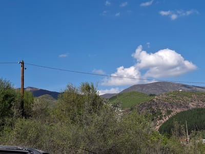 A vibrant, daytime, wide-angle shot captures a rural mountain landscape in Zverino, Bulgaria, under a bright blue sky dotted with scattered white cumulus clouds. The scene is dominated by a clear horizon where the sky meets multiple layers of hills and mountains.

In the foreground, dense deciduous bushes and trees with fresh, light green spring foliage fill the lower and mid-sections of the frame, some exhibiting small white blossoms. A prominent wooden utility pole, equipped with ceramic insulators and carrying electrical wires, stands on the left side, with its wires extending horizontally across the midground. Part of a dark-colored vehicle, possibly a car hood or roof edge, is visible at the very bottom left corner.

The midground features undulating hills covered in a mix of green trees and bushes, while further back, some slopes show sparser, darker vegetation and rocky outcrops. In the distance, higher mountain peaks appear rugged and dark. On one of the more distant hills to the right, a communications tower or antenna structure is discernible. Further to the right, nestled among the trees, a few houses with distinct red or orange tiled roofs and white walls are faintly visible, suggesting a small settlement.

The weather appears clear and sunny, indicating good visibility. There is no discernible human activity or specific event taking place, and no visible text is present in the image. The overall impression is one of natural tranquility in a mountainous, rural setting.
