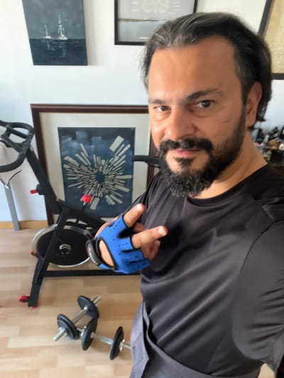 This media file shows a man indoors, appearing to be in a home gym. The man has dark hair and a beard, and is wearing a black t-shirt and blue weightlifting gloves. He is holding up his right hand, gloved, and pointing with his index finger. In the background, there is a stationary exercise bike and two dumbbells on the floor. A framed abstract artwork hangs on the wall behind the bike, and another framed picture is visible on the left. The lighting suggests it is daytime. The scene implies the man is either about to, or has just finished, a workout.