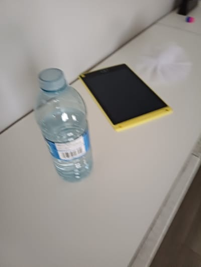 The image shows a clear plastic water bottle with a blue cap and a label on a white surface. Next to the water bottle is a black tablet device with a yellow frame. To the right of the tablet, there is a blurred white object. The scene appears to be indoors, possibly on a desk or table, with a white wall in the background. There are no people visible in the image. The overall lighting is soft. The image is taken from a slightly elevated angle, looking down at the objects. There is no information available about the specific location in Morden, Canada.