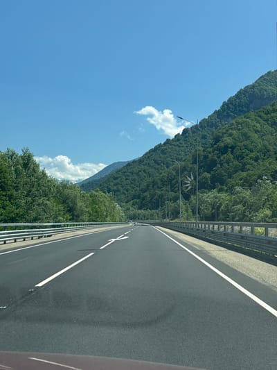 The image shows a highway with guardrails on both sides, surrounded by lush green trees and mountains under a clear blue sky. The highway has clear white lane markings and appears to be a multi-lane road. There are streetlights visible along the side of the road. In the distance, the road curves slightly as it enters a mountainous area. A unique decorative structure, resembling a large pinwheel or flower, is visible on a pole among the trees on the right side of the road. The scene suggests a scenic drive through a natural landscape, possibly in a rural or mountainous region of Russia, given the mention of Chvizhepse.