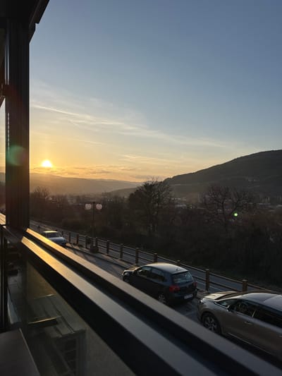 The image displays an outdoor panorama at either sunrise or sunset within Dimitrovgrad Municipality, Serbia, viewed from an elevated vantage point behind a dark, possibly metallic, architectural frame. The sun, a bright yellow orb, is low on the left horizon, casting strong golden light and long shadows across the scene. The sky transitions from light blue at the zenith to orange and yellow at the horizon, with sparse, wispy clouds.

In the midground, a paved road is visible, bordered by a fence with wooden posts. Parked on the right shoulder is a dark gray Volkswagen Golf hatchback with a Serbian license plate "BG 147 AA" and a beige or champagne-colored sedan. A silver or light-colored sedan is partially visible parked on the left shoulder of the road. A dual-globed street lamp stands by the roadside. Beyond the road, a valley containing indistinct settlement structures leads to multiple layers of hills and mountains, predominantly covered with bare deciduous trees, indicating a colder season.

The immediate foreground consists of dark, angular elements of a window or balcony frame, with reflective surfaces showing indistinct reflections of an interior space. No individuals are present, and no actions or interactions are depicted.