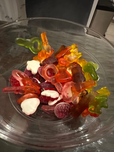 The image shows a close-up, overhead view of a clear glass bowl filled with an assortment of colorful gummy candies. The candies are varied in shape and color, including gummy bears in green and yellow, red and orange ring-shaped candies, white and red candies resembling lips or dental objects, and dark red, raspberry-shaped gummies. The bowl is placed on a dark, textured surface, possibly a wooden table. The background is dimly lit and out of focus, showing hints of furniture and wall textures, suggesting an indoor setting. There is no information in the image to suggest a location in Recklinghausen, Germany.