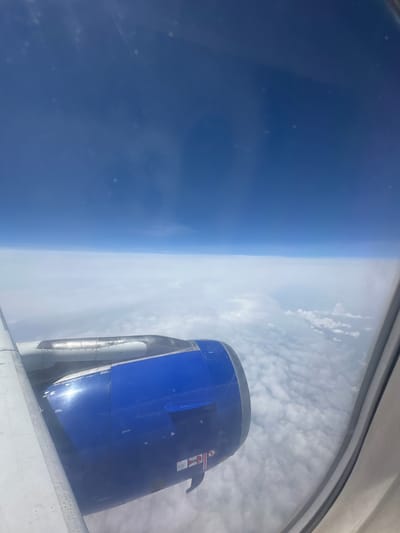 The image, taken from an airplane window, shows a view of the Earth's atmosphere from high altitude. The foreground features a portion of an airplane's blue engine nacelle and part of the wing. The background is dominated by a vast expanse of white clouds, layered beneath a deep blue sky. There is no indication of the city of Viehhausen, Austria, in this image.