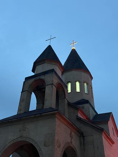 The image shows a low-angle view of a church structure against a clear blue sky. The church is constructed of gray stone with reddish-brown accents. It features two prominent towers with conical roofs, each topped with a cross. The tower on the left has a simple cross, while the tower on the right has a more ornate, glowing cross. The structure includes architectural details such as arched openings, decorative crosses, and geometric patterns on the stone surface. The scene is likely in Novomikhaylovsky, Russia, given the prompt's reference to that location.