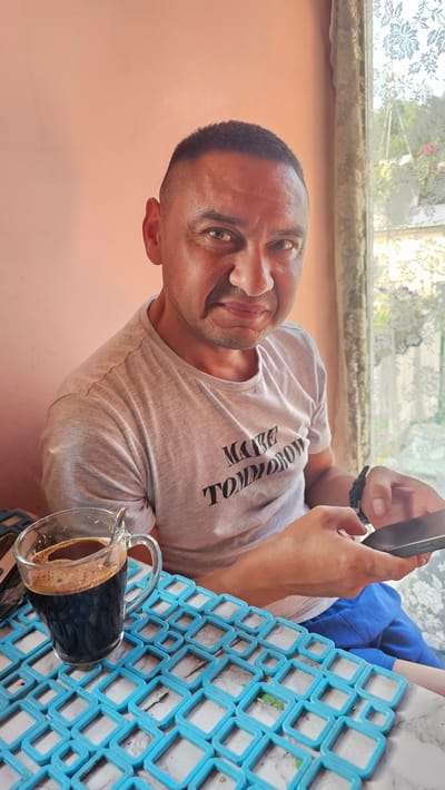 A middle-aged man with a buzz cut sits at a table with a cup of dark coffee to his left. He is wearing a light grey t-shirt with the words "MAKE TOMORROW" printed on it in black, bold lettering. The man is looking at the camera with a slightly displeased expression and holding a smartphone in his right hand. To the right of the man is a window with a lace curtain. The table is covered with a light-colored marble-patterned surface and a blue, square-patterned placemat. The background wall is a pale pink. The setting appears to be indoors, possibly a home or cafe. The image was taken in Nemėžis, Lithuania.