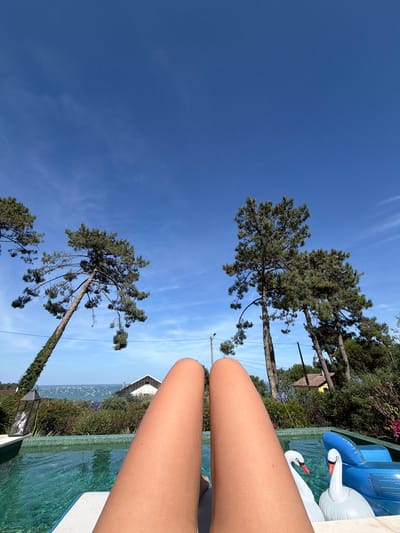 A person's legs are visible in the foreground, lying on a pool lounger. The legs are tanned and positioned so that the view is directed towards the horizon. In the mid-ground, a clear blue swimming pool is filled with water, with two inflatable swan pool toys and a blue inflatable lounge chair floating on the surface. Beyond the pool, a lush green garden with various plants and flowers is visible. Several tall pine trees with green needles are scattered throughout the scene, some leaning towards the sky. In the distance, several white houses with red-tiled roofs are nestled amongst the trees, and further still, the ocean is visible with numerous boats anchored in the water. The sky above is a clear, vibrant blue with a few wispy white clouds. The image appears to be taken on a sunny day in Lège-Cap-Ferret, France, likely during the summer.