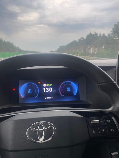 The image shows the interior of a car dashboard, focusing on the digital display and a portion of the steering wheel. The car is traveling at 130 km/h, indicated on the central display. To the left of the speed reading, a fuel gauge shows 340 km remaining. To the right, the RPM is displayed as 2400. The odometer shows 290,622 km. A warning light, possibly for the exhaust system, is illuminated on the left side of the display. The background shows a cloudy sky and a dense line of trees, suggesting a highway or rural road. The steering wheel features the Toyota logo. The image was captured in Tarnawa Rzepińska, Poland.
