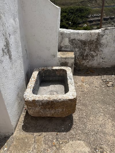 The image shows a close-up view of an old, rectangular stone trough situated on a concrete ground surface. The trough is worn and weathered, with visible signs of age and exposure to the elements. It appears to be made of rough-hewn stone, with a rough, irregular texture along its edges. The interior of the trough is darker and appears to be filled with dirt or debris. To the left of the trough is a white, stucco-covered wall with visible texture and some areas of discoloration or staining. Behind the trough, a series of rough, concrete steps ascend. Further in the background, a weathered white wall continues, with a rough, mottled texture. The ground surface around the trough is a mix of concrete and gravel, with some loose stones and debris scattered about. A shadow is cast by the trough onto the ground. In the upper right portion of the image, a wooden post is visible, and beyond that, a hillside covered with vegetation and some rocky outcrops can be seen. The scene suggests an outdoor setting, possibly in a rural or historical area. The question states the location is Colares, Portugal, but this cannot be visually confirmed from the image alone.