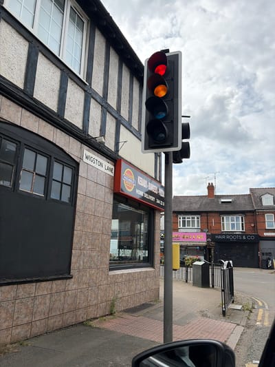 The image depicts a street scene in Leicester, United Kingdom, specifically at the intersection of Wigston Lane and Aylestone Road. A traffic light is showing a red and amber signal. Adjacent to the traffic light, a building has a sign for "Pizzas, Kebabs, Burgers, Pizza" and "Home Delivery 244 2288". To the right of this building is a shop called "Hair Roots & Co, Est. 2014", which sits on Aylestone Road. Part of a vehicle is visible on the right side of the image. The sky is overcast with clouds.