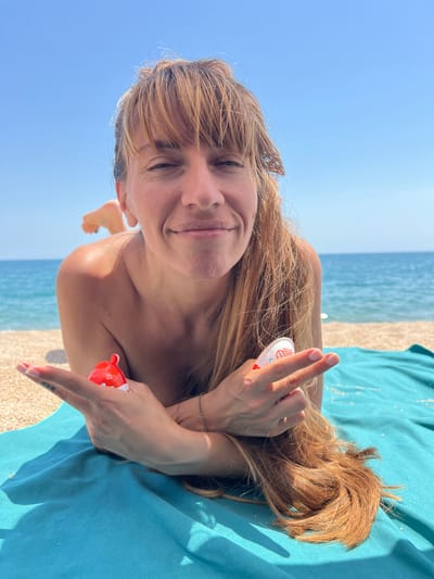 A woman is lying on a blue towel on a beach with the sea and sky in the background. She is looking at the camera and smiling, with her hands outstretched. She is holding a red and white Kinder Surprise egg in each hand. The location is the beach in Sant Pol de Mar, Spain.
