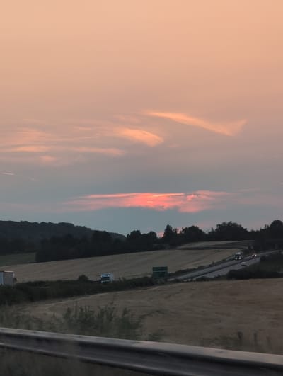 The image shows a landscape scene with a highway and fields under a dusky sky. In the foreground, a metal guardrail is visible. Beyond the guardrail, there are harvested fields, some with dark green bushes and trees interspersed. A highway curves through the landscape, with a truck and a car visible on it, their headlights on. A green highway sign is also present. In the background, rolling hills covered in trees form the horizon line. The sky is filled with soft hues of pink and orange, indicative of a sunset or sunrise, with some wispy clouds. The overall impression is a rural or semi-rural setting at twilight. The location is identified as being in the vicinity of Domgermain, France.