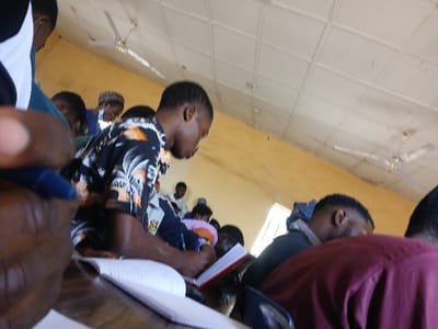 This image depicts an indoor scene, likely a classroom or lecture hall, situated in Ugi Majowel, Nigeria. The perspective is from behind and slightly to the left of a seated individual, showing their hand holding a blue pen over an open notebook on a dark desk.
In the mid-ground, a male individual with dark skin and short, dark hair is seated, facing right and downward, focused on a red notebook. He is wearing a black short-sleeved shirt with a multi-colored abstract pattern featuring orange and white elements. He holds a black pen in his right hand, poised over the notebook.
To his right and slightly behind him, another male individual with dark hair is visible, wearing a dark blue or black top with a patterned hood, also looking downward. The back of a third person, wearing a maroon or dark red garment, is visible in the immediate foreground on the right.
In the background, several other individuals are seated, partially obscured, appearing engaged in similar activities. One person in the deeper background wears a light blue face mask. The room features pale yellow walls and a light-colored, paneled ceiling from which two white, two-blade ceiling fans are suspended. A window or light source is visible in the upper right background. The overall activity suggests a learning or academic setting where individuals are focused on writing or studying.