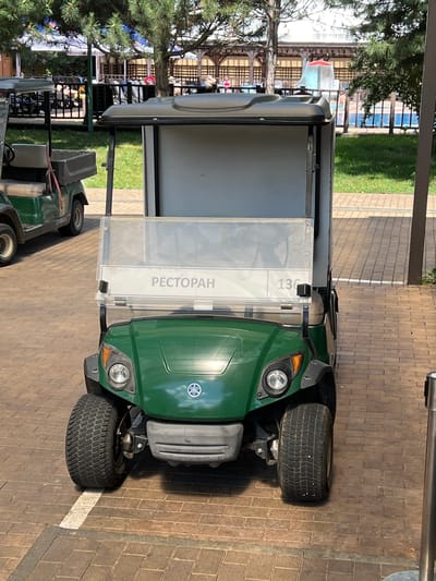 This image shows a green Yamaha golf cart parked on a brick-paved walkway. The golf cart has a clear windshield with a sign that reads "РЕСТОРАН" (Restaurant) in Russian and the number "136". There is another golf cart visible in the background to the left. Behind the golf carts, there are trees, a fence, and what appears to be a swimming pool area with people in the distance. The environment suggests an outdoor recreational or resort location. The specific location in the city of Plast, Univska, Russia, is not explicitly discernible from the image itself, but the presence of a golf cart and pool area suggests a leisure facility.
