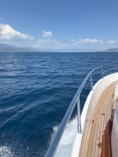 This media file captures a view from the deck of a boat moving through the ocean. The camera is angled downwards towards the water, with the railing of the boat prominently in the foreground. The water is a deep blue with visible ripples and some white spray along the side of the boat, indicating movement. The deck of the boat is made of polished wooden planks, some of which appear wet.
In the distance, a hazy coastline is visible, featuring hills or mountains. There are also several white, vertical structures that resemble wind turbines on the distant land. The sky is a clear, bright blue with scattered white clouds, suggesting a sunny day.
The overall impression is one of a serene journey at sea, likely during daylight hours, given the bright sunlight and clear blue sky. The scene is located off the coast of Patras, Greece, as indicated by the specific details of the landscape and potential wind farms. No people are visible in the frame, and there is no visible text.