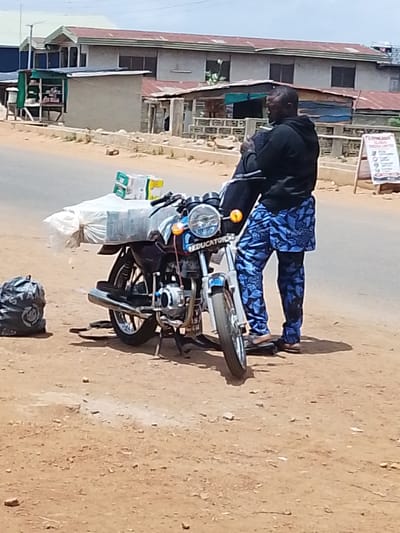 A man stands next to a motorcycle laden with goods in Iseyin, Nigeria. The man, wearing a black hoodie and blue patterned trousers, appears to be holding a dark bag or garment. The motorcycle has a large bundle wrapped in white plastic and several boxes stacked on its rear carrier. To the left of the motorcycle, a black plastic bag lies on the ground.

The scene is set on a dirt road next to a paved street. In the background, there are buildings with corrugated iron roofs and some smaller structures that appear to be shops or stalls. One stall, with a green awning, displays various items on shelves. A white sign with text is visible on the right side of the image, possibly advertising services or products.

The lighting suggests it is daytime, with bright sunlight casting shadows. The overall atmosphere is that of a typical roadside scene in a town or city. There is no clear indication of the specific time of day beyond it being daylight. The weather appears to be dry and sunny. The visible text on the motorcycle's fender reads "EDUCATOR." The black plastic bag on the ground has some white lettering on it, but it is not entirely legible. The white sign in the background has text that includes "GLOBAL NIGERIA LIMITED" and "POS."