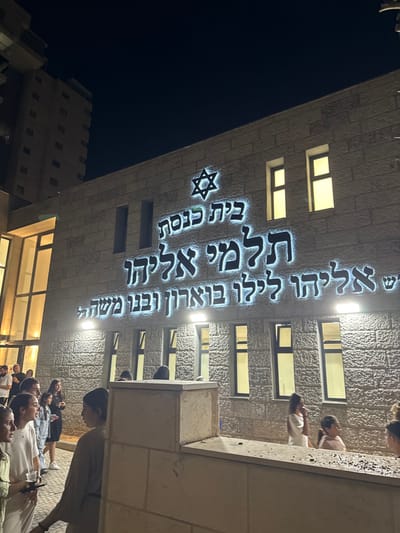 The image displays the exterior of the Talmei Eliyahu Synagogue at night, situated in the Remez neighborhood of Beer Yaakov, Israel. The building features a light-colored, stone-block facade. Prominently illuminated on the front is a large Hebrew sign that reads "בית כנסת תלמי אליהו" (Talmei Eliyahu Synagogue), followed by "אליהו לילי בוארון ובני משה" (Eliyahu Lily Boaron and sons Moshe), with a glowing Star of David emblem positioned above the text. Multiple rectangular windows, some emitting light, are visible across the building's face.

A crowd of approximately fifteen individuals, including adults and children, is gathered outside the synagogue, primarily on the lower left and right portions of the frame. They appear to be standing and conversing. One adult woman on the far left holds a clear cup, and another holds a smartphone. A low, light-colored concrete barrier or wall runs horizontally across the foreground. In the upper left background, a multi-story building with multiple balconies is partially visible against a dark, clear night sky.