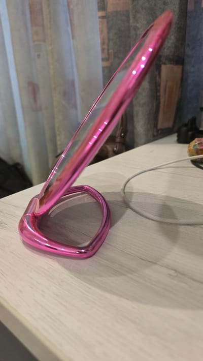 The image shows a close-up, tilted view of a pair of pink metallic glasses. The glasses have a distinct metallic sheen and are positioned on a light-colored wooden surface. In the background, there is a blurry patterned wallpaper with brick-like designs and what appears to be a light gray cable. A brown handbag is also partially visible in the background. The lighting suggests an indoor setting, possibly during the daytime. There is no visible text in the image. The focus is on the texture and reflective quality of the glasses, which appear to be the primary subject. The scene is static, with no apparent activity taking place. The location cues are general, with the wooden surface and wallpaper not providing specific indicators of a unique setting.