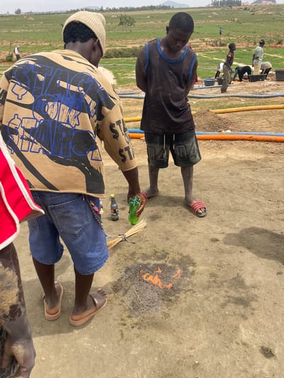 Two men stand outdoors in Maiyanga, Nigeria, near a small fire on a dirt surface. One man wears a black tank top and dark shorts with red sandals, facing away from the camera. The other man, closer to the camera, wears a tan and black patterned shirt, blue shorts, and a tan beanie, holding a green bottle. A broom and a dark bottle lie on the ground next to the fire. In the background, a group of people are working near blue and orange pipes, set against a grassy landscape.