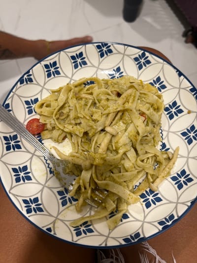 A plate of tagliatelle pasta with pesto sauce is presented, speckled with small tomato pieces. A silver fork rests on the plate, partially obscured by the pasta. The plate features a white base with a blue and white geometric pattern resembling stylized flowers. The plate is positioned on a white tiled floor, with portions of a person's leg and foot visible at the edges of the frame.