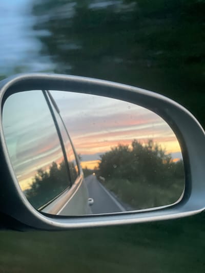 The image shows a reflection in a car's side mirror, capturing a sunset over a rural road. The sky displays a gradient of soft oranges, pinks, and pale blues, with wispy clouds stretching horizontally. Silhouetted trees line the sides of the road, which recedes into the distance. The car's silver exterior is visible in the foreground of the reflection. The surrounding environment is dark green and appears to be foliage, suggesting a natural setting outside of a city. There are no people visible in the image.